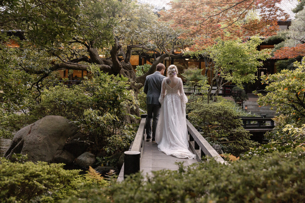 The bride and groom walk across a small wooden bridge into a Japanese garden as the maples turn red.