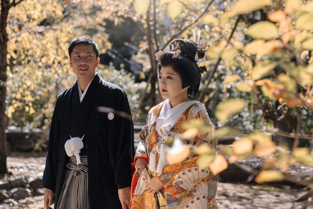 A bride in a gold and orange iro-uchikake and traditional hair ornaments walks beside the groom in black montsuki and hakama among autumn leaves.