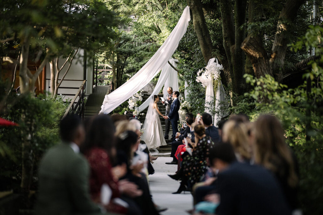 Guests line a garden aisle at Jinya as the bride and groom stand beneath white drapes for their ceremony at a ryokan wedding in Japan.