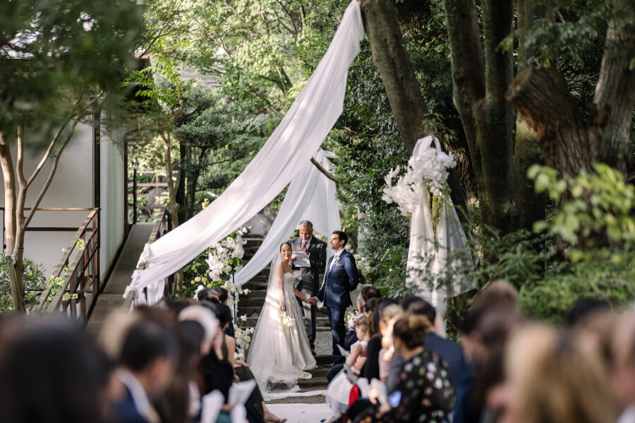 The bride and groom stand before their officiant beneath white drapery hung through the trees for their garden ceremony at Jinya, a ryokan wedding in Japan.