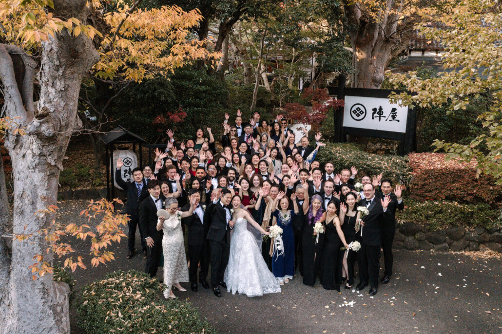 The whole wedding party waves up at the camera beneath the Jinya sign and a canopy of autumn leaves.