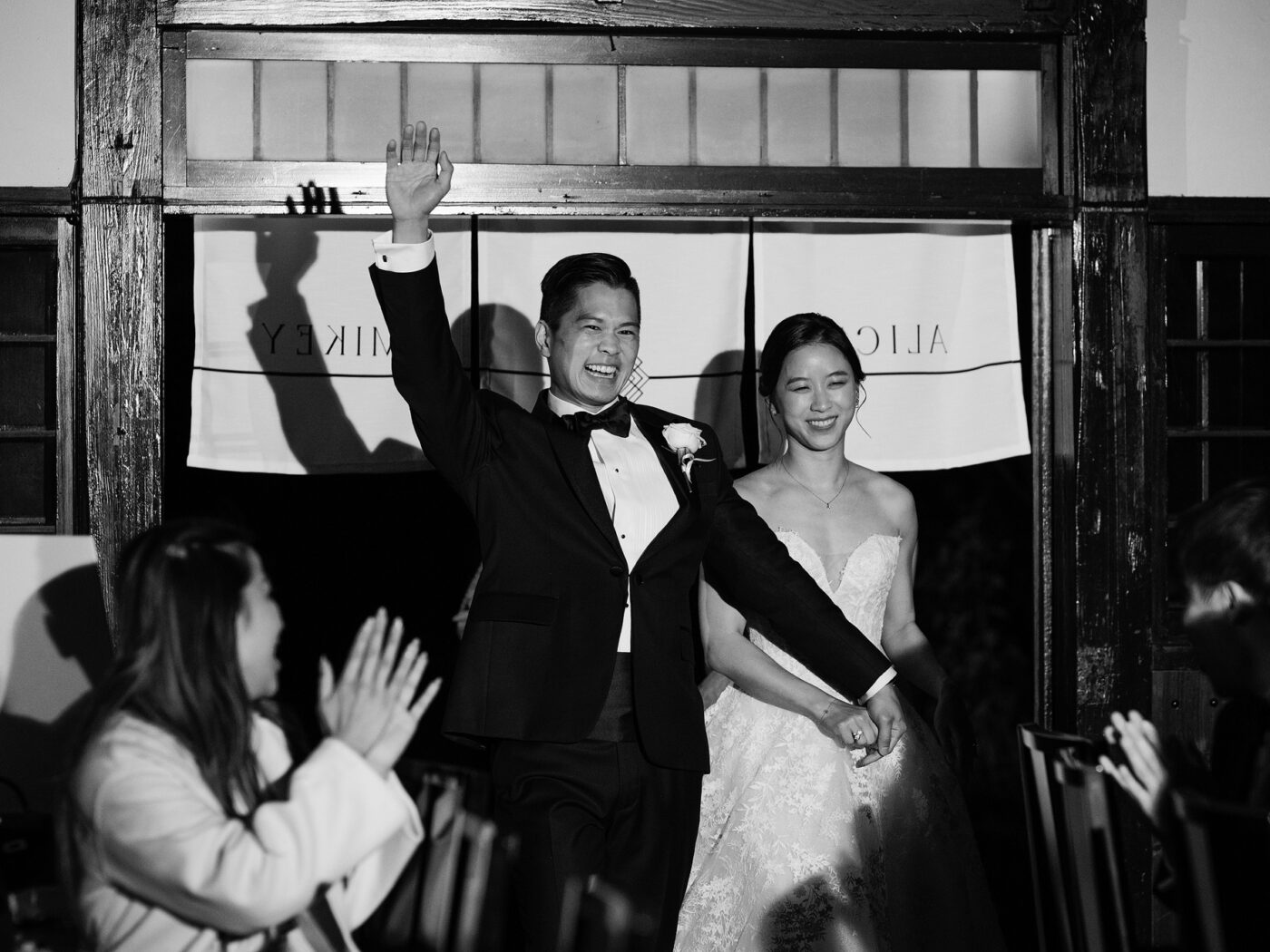 In black and white, the groom throws his arm in the air as he and the bride walk through a noren curtain into their wedding reception at Jinya.