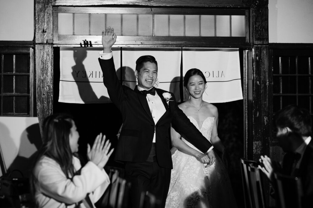 In black and white, the groom throws his arm in the air as he and the bride walk through a noren curtain into their wedding reception at Jinya.