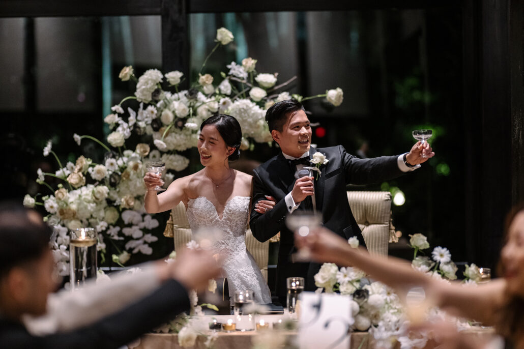The bride and groom raise their glasses from a table banked with white flowers as guests toast them at their wedding reception at Jinya.