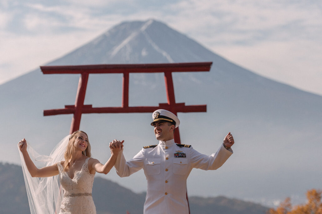 Arms flung wide and laughing, the couple celebrate beneath the red torii with the mountain behind, their Mt Fuji wedding portraits.