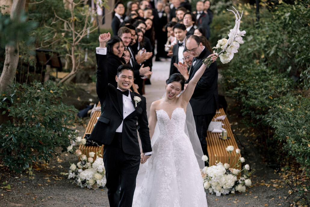 The bride raises her bouquet and the groom his fist as they reach the end of the aisle, laughing.