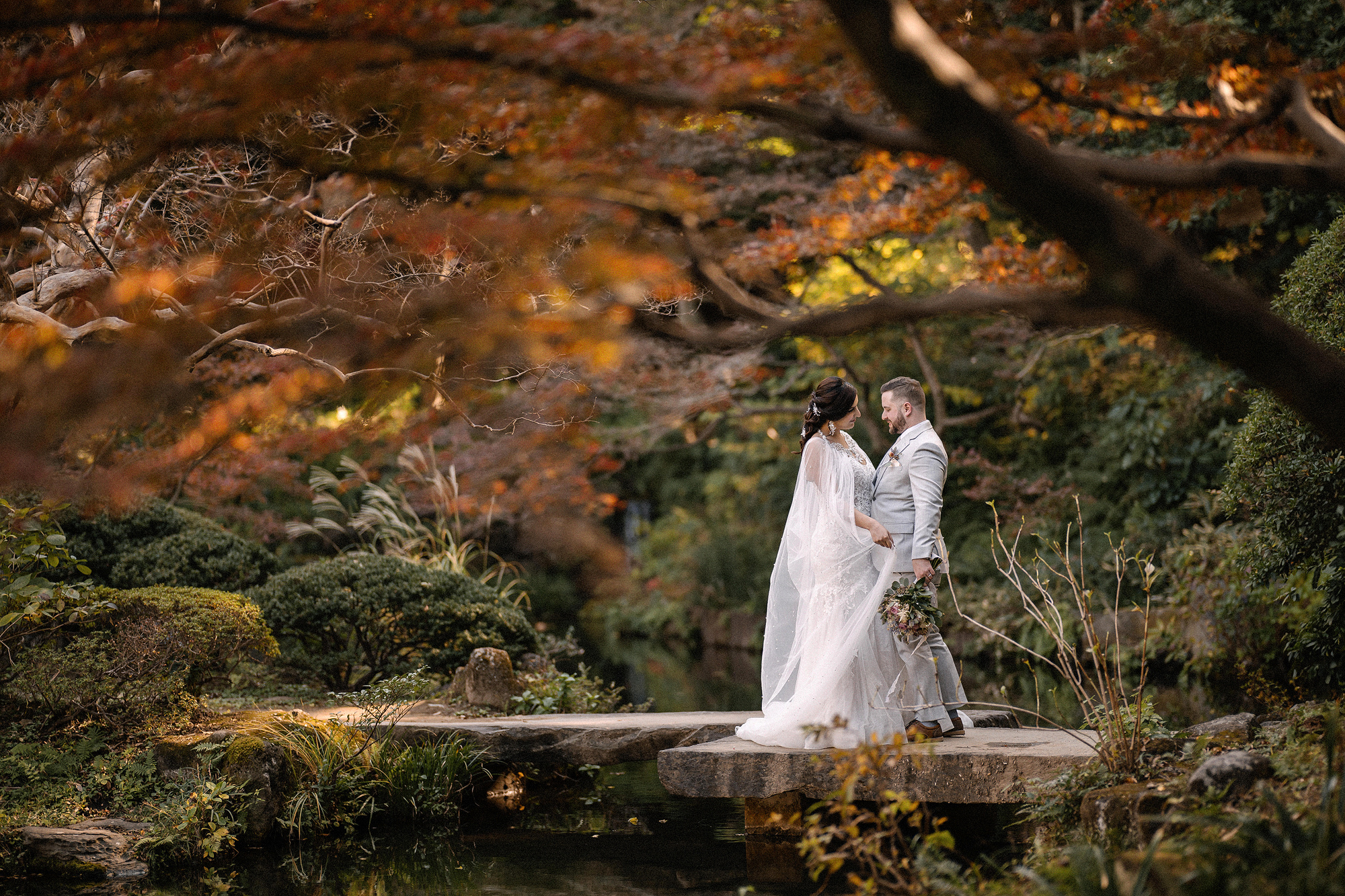 The bride and groom stand close on a stone slab bridge across the pond, maples overhead, in Kansen-en, a small garden in Shinjuku.