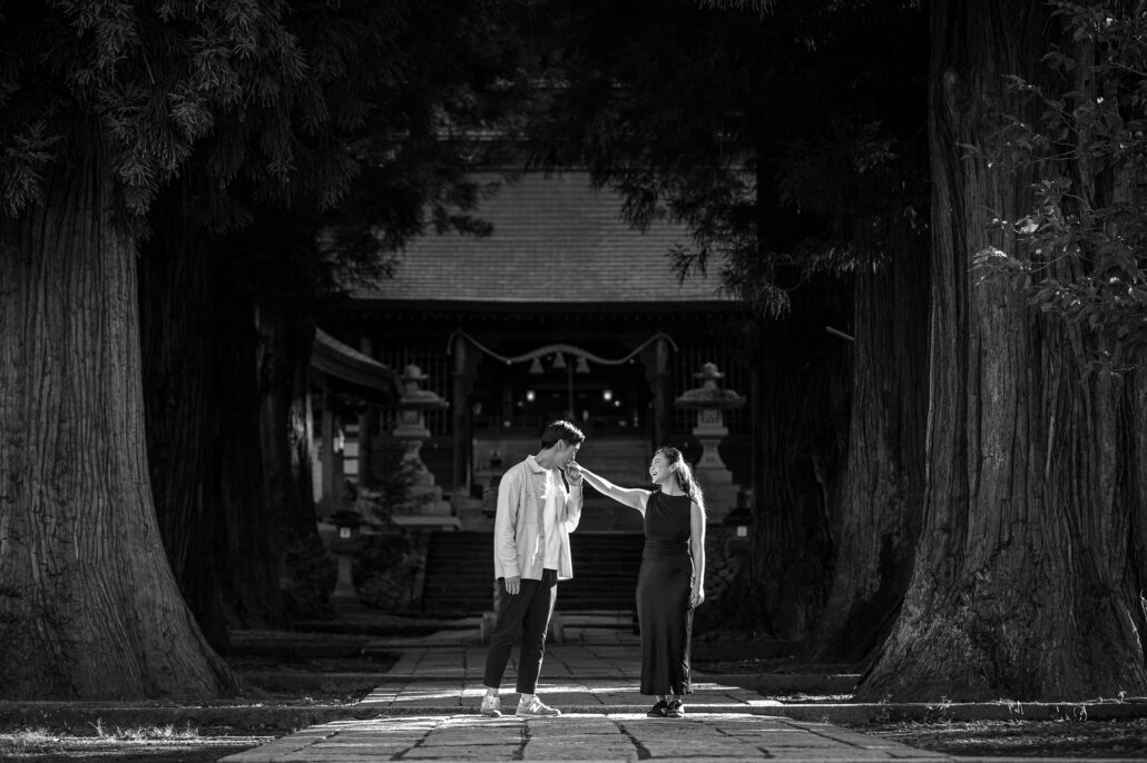 In black and white, he kisses her hand on the stone path at Kawaguchi Asama Shrine, giant old cedar trunks rising on both sides.