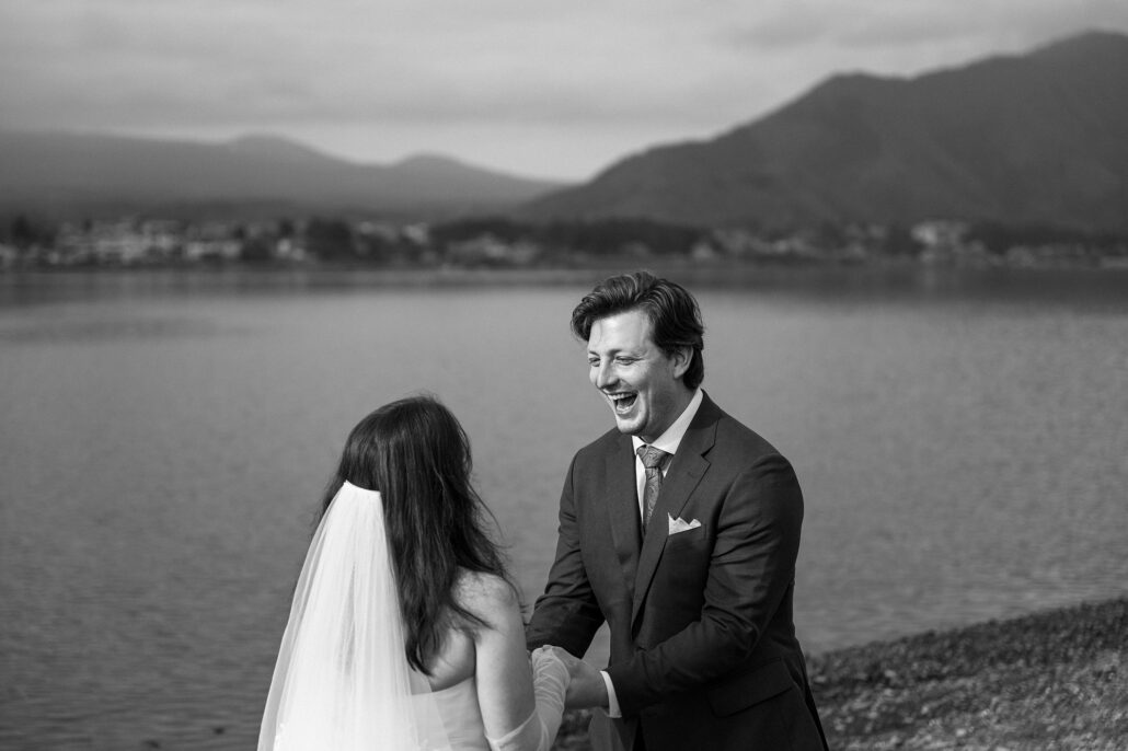 In black and white, the groom laughs as he holds the bride's hands by the water during their elopement vows at Lake Kawaguchiko.