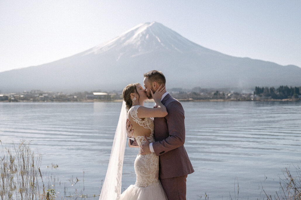 The bride and groom kiss by the water with Mt Fuji sharp against a winter sky, just the two of them at their Kawaguchiko elopement.