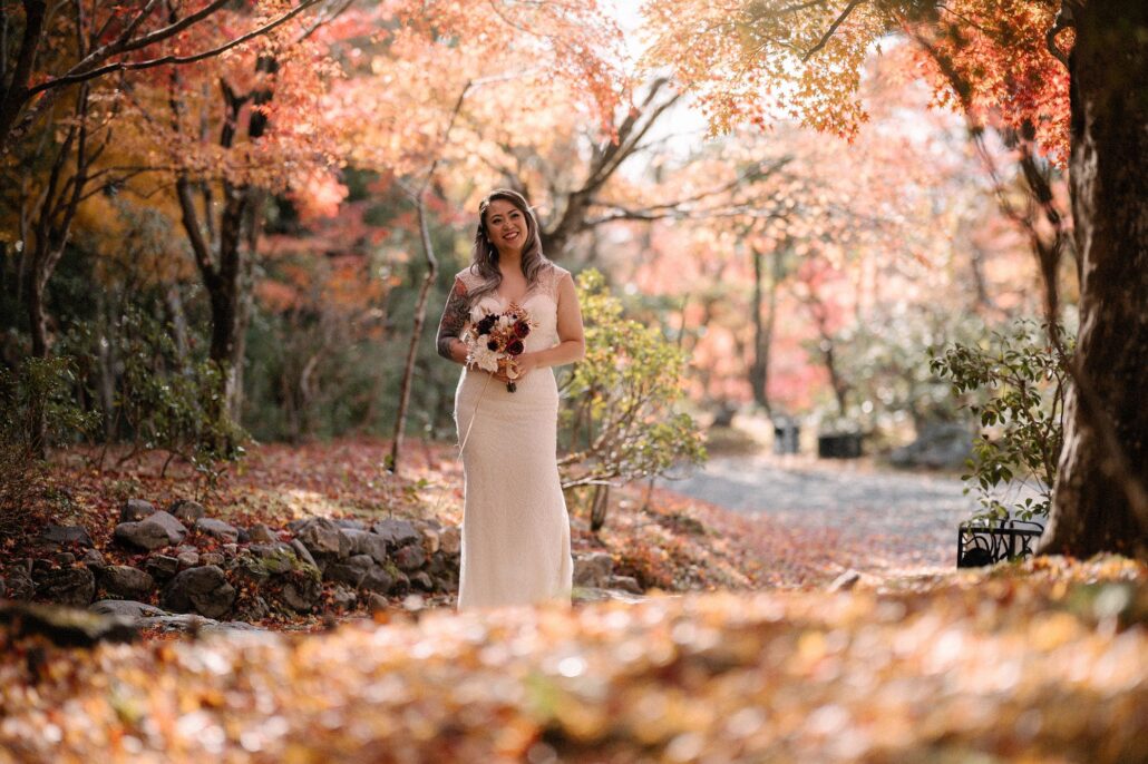 The bride walks down a garden path carpeted with fallen red leaves toward the groom.