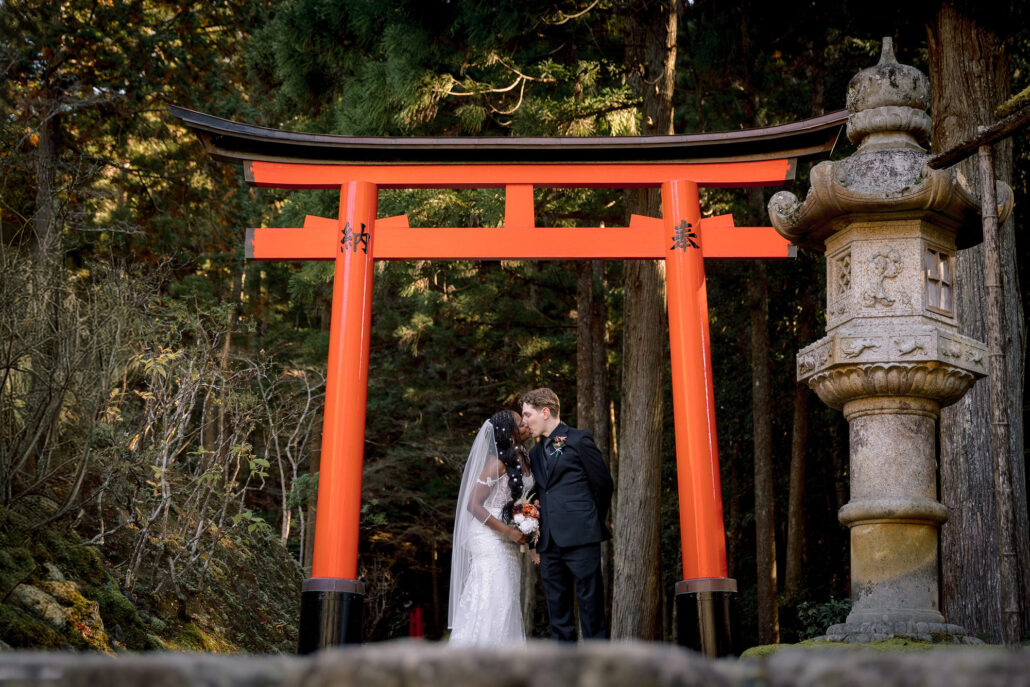 The bride and groom kiss beneath a vermilion torii beside a stone lantern in a private garden in Kyoto.