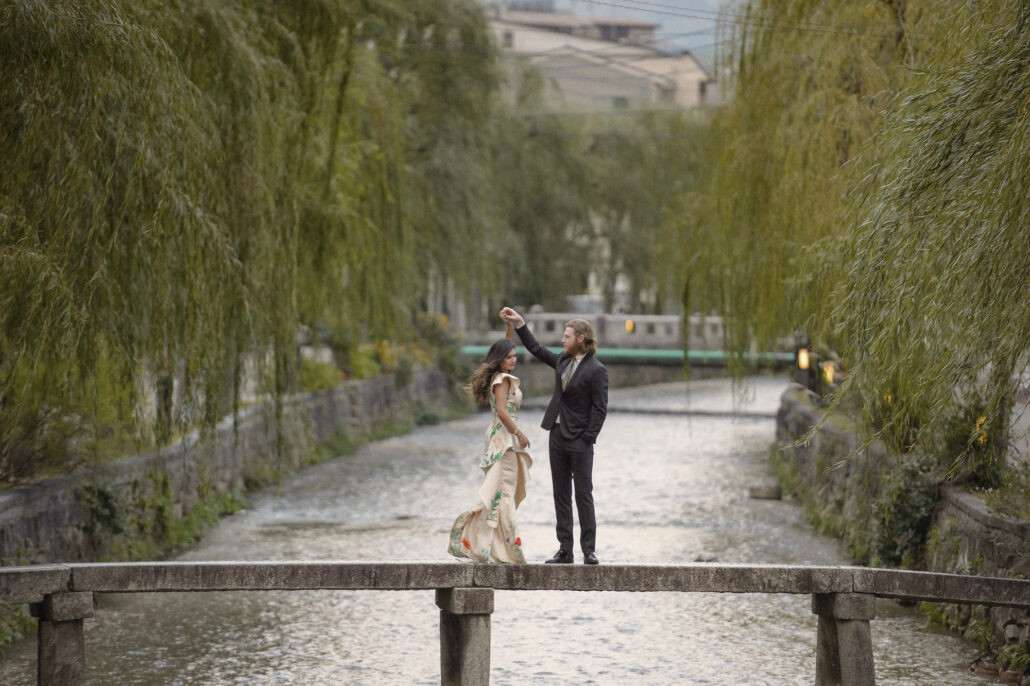 A Kyoto elopement on a narrow stone bridge over the willow-lined Shirakawa, the groom raising the bride's hand.