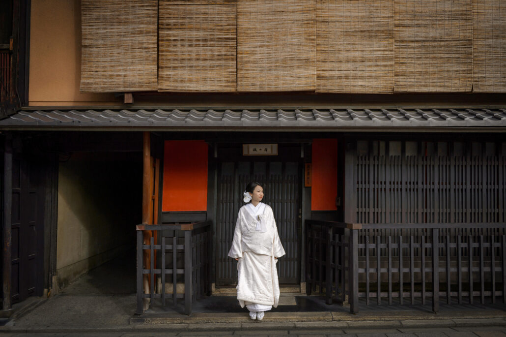 A bride in a white wedding kimono at the doorway of an old wooden machiya on a quiet Kyoto street, bamboo blinds above her.