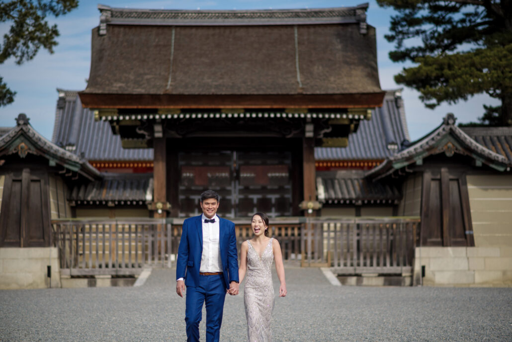 A couple walk hand in hand across the gravel before a great wooden gate in Kyoto on a quiet morning, pines and a clear blue sky behind.
