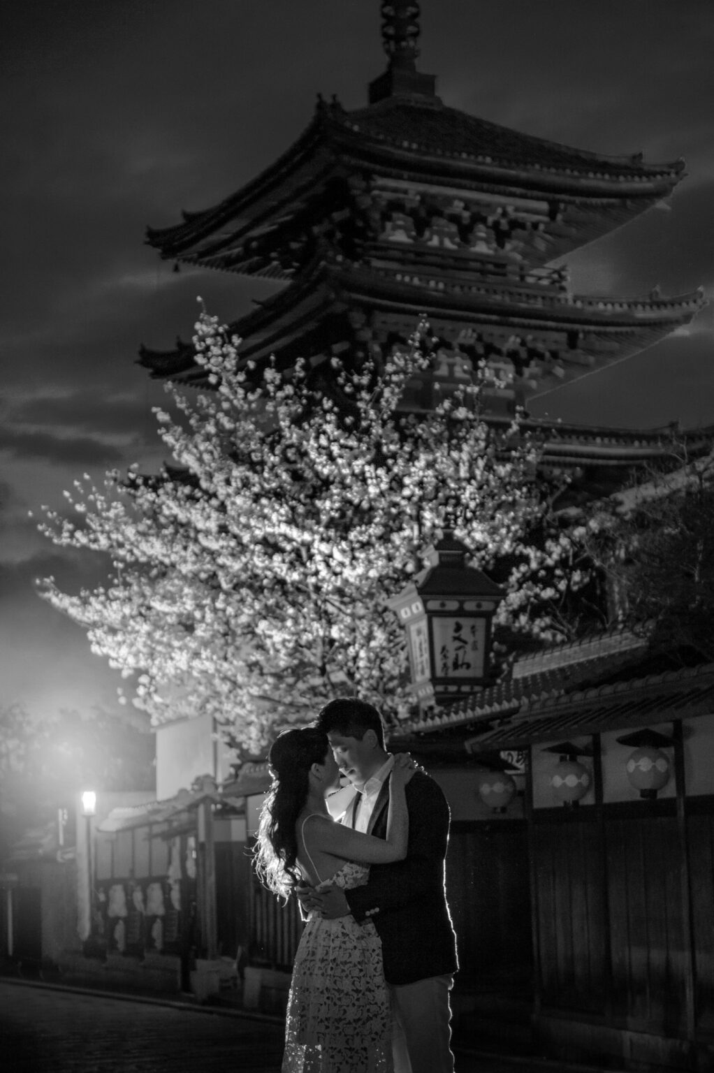 In black and white, a couple kiss beneath Yasaka Pagoda in Kyoto at night, cherry blossom lit above the quiet lane.