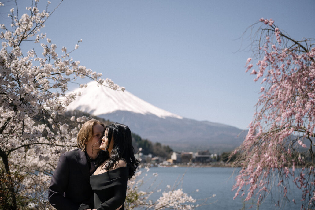 The couple kiss on the shore of Lake Kawaguchiko beneath weeping cherry, Mt Fuji white with snow across the water.