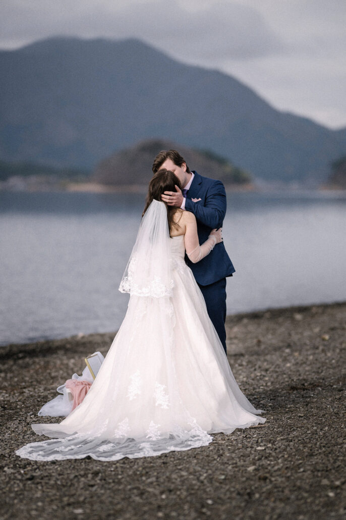 The couple embrace on the lakeshore with a small wooded island and mountains behind, a Lake Kawaguchiko elopement in spring.