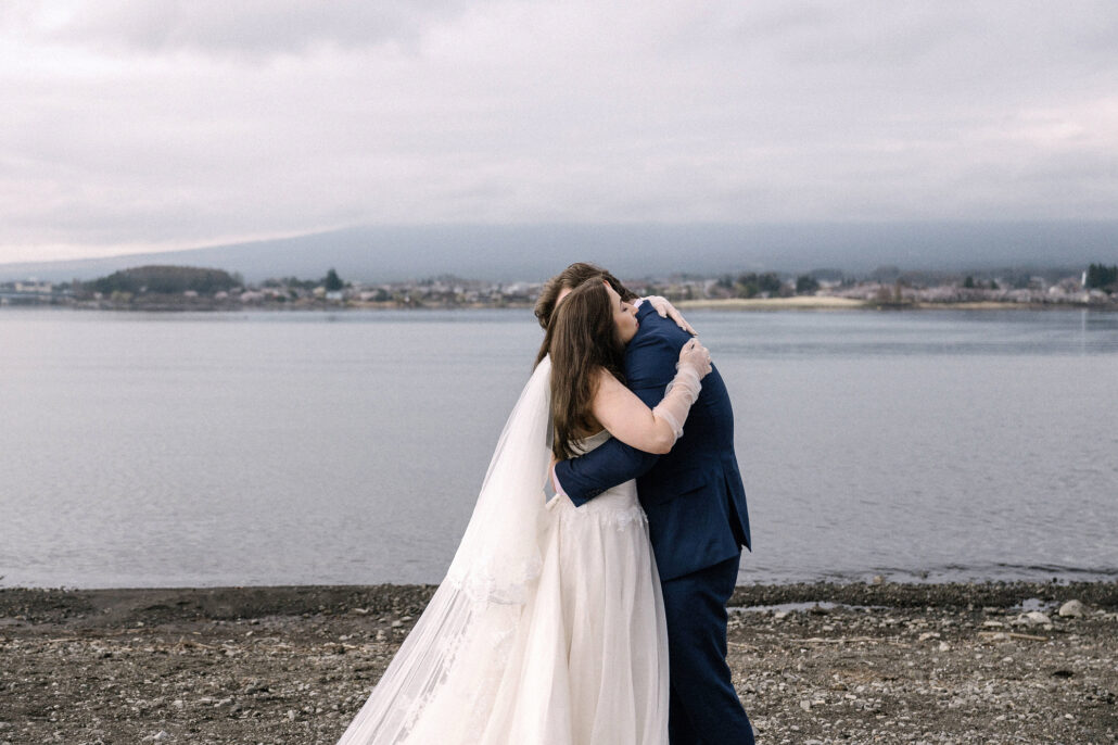 The bride and groom embrace on the stony shore of Lake Kawaguchi while Mt Fuji stays hidden in cloud on their Fuji Five Lakes elopement.