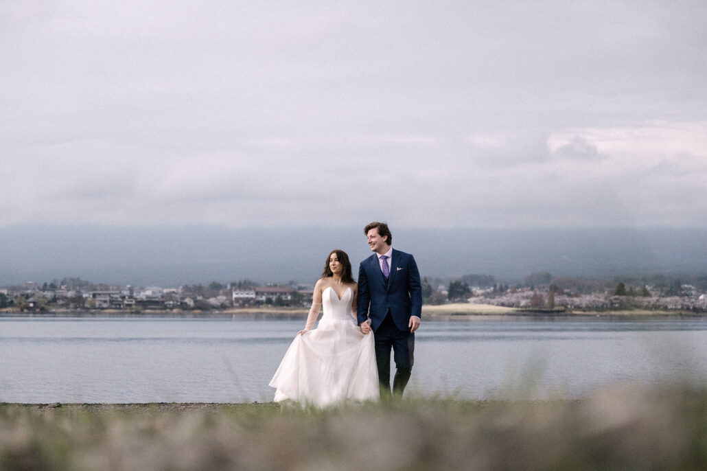 The bride lifts her skirt as she walks with the groom along the shore of Lake Kawaguchi, low cloud on the far hills, a Japan elopement.