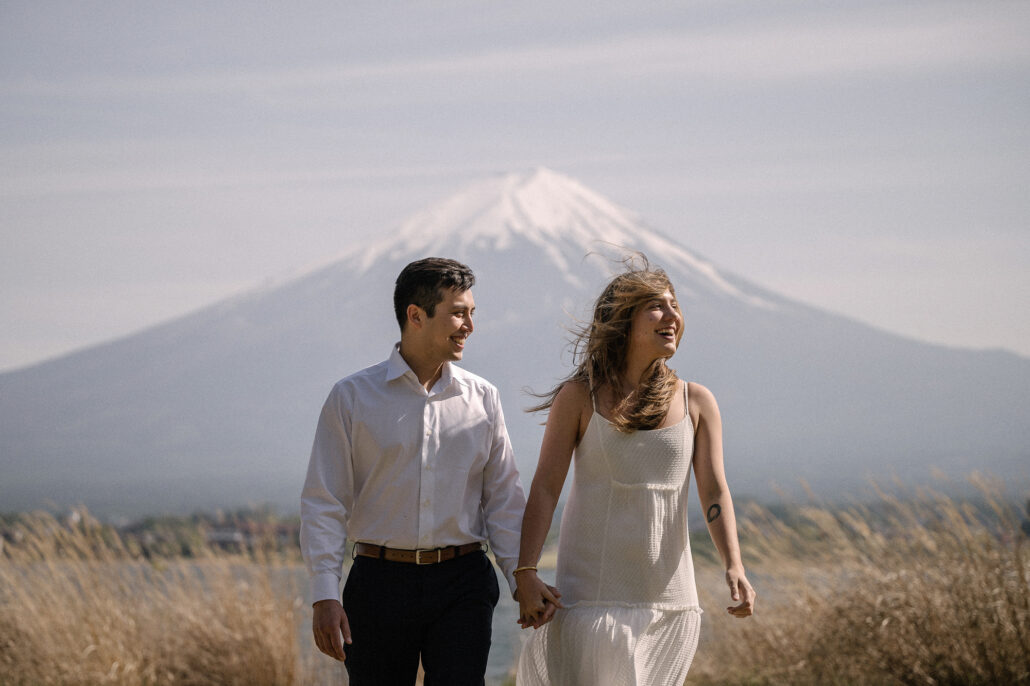 Hand in hand, the couple walks through dry gold grass by the lake, her hair lifting in the wind, Mt Fuji in spring behind them.