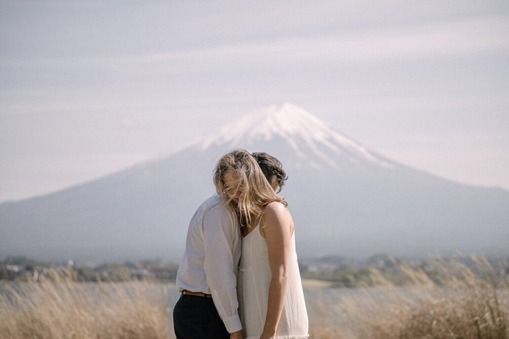 Her hair blowing across her face, she leans into his embrace on the lakeshore at Kawaguchiko, Mt Fuji's snowy summit above.