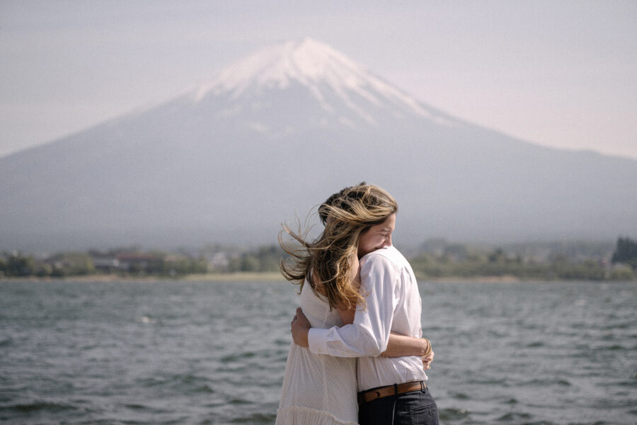 The couple holds each other at the windy water's edge, her hair blowing, after a Lake Kawaguchiko proposal with Mt Fuji behind.