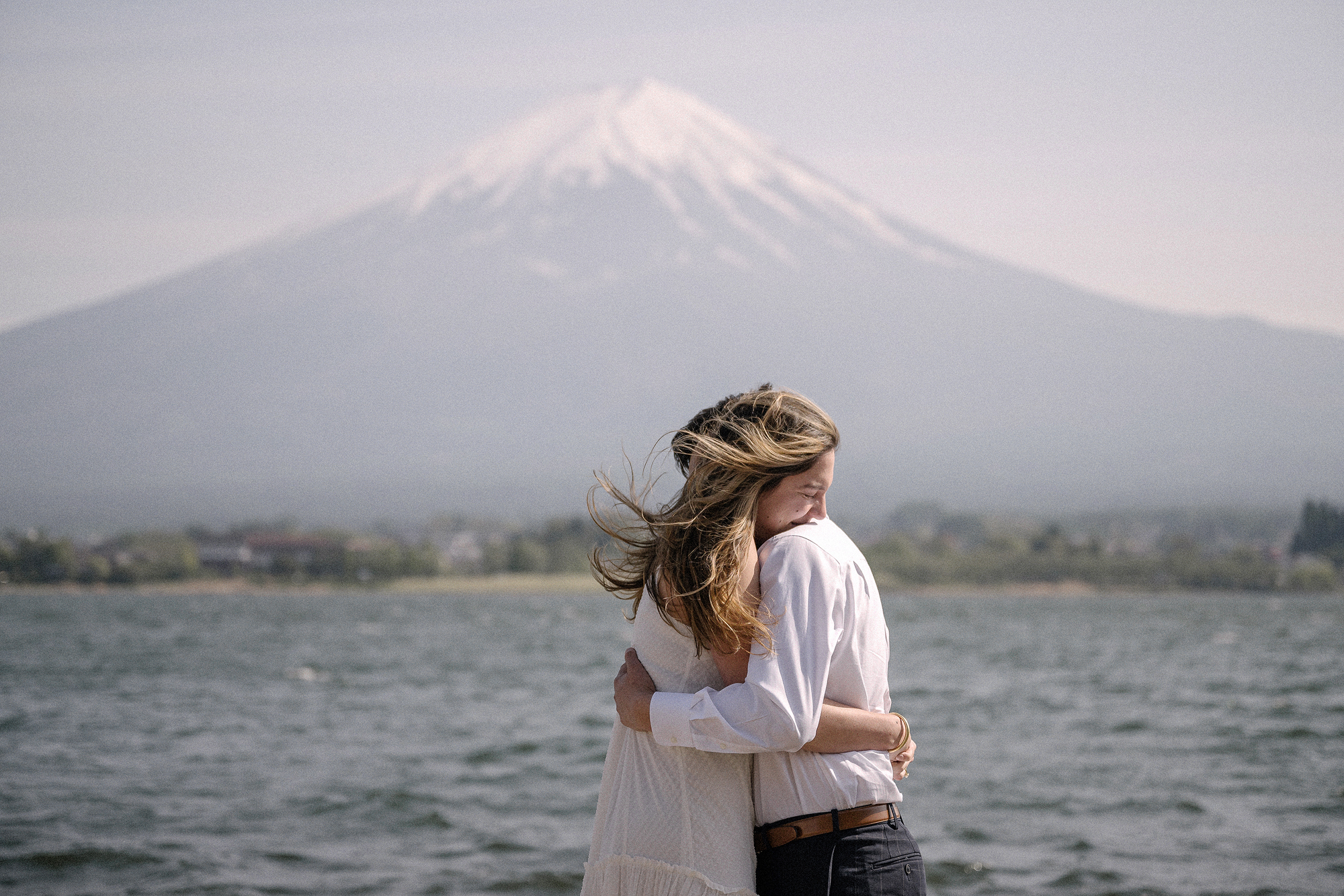The couple holds each other at the windy water's edge, her hair blowing, after a Lake Kawaguchiko proposal with Mt Fuji behind.