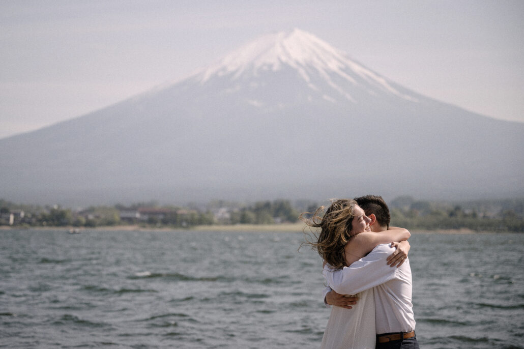 Wind lifts her hair as the couple hugs at the edge of the choppy lake, a surprise proposal at Kawaguchiko with Mt Fuji behind.