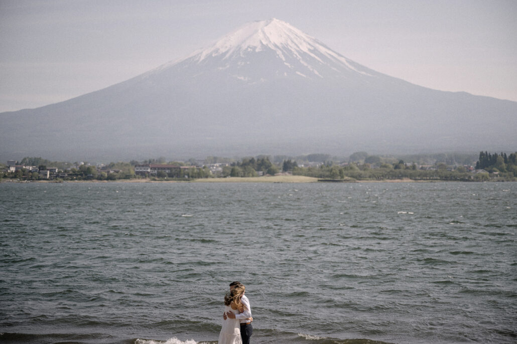 A wide view of the newly engaged couple holding each other at the edge of Lake Kawaguchi, Mt Fuji rising over the far shore.