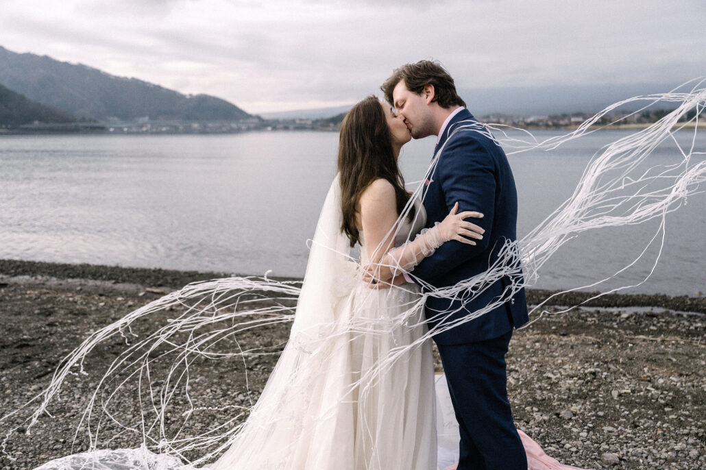 White streamers fly around the bride and groom as they kiss on the lakeshore after their vows, an elopement at Lake Kawaguchiko.