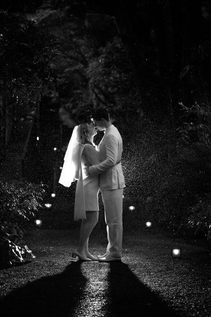 The couple embrace on a wet garden path lined with small lanterns, long shadows behind them on a rainy elopement night in Japan.