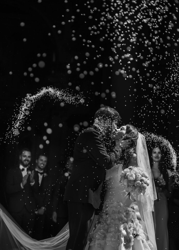 In black and white, the bride and groom kiss as their guests throw rice over them in a glittering arc, at a destination wedding in Lecce, Italy.