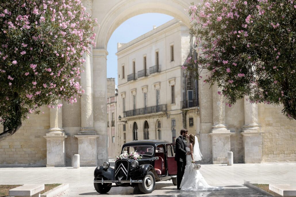The bride and groom kiss beside a vintage black car under a stone arch and flowering trees in Lecce, Italy.