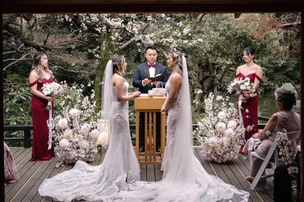 Two brides stand at a wooden lectern for their LGBTQ wedding in Japan, bridesmaids in burgundy beside them and real cherry blossom overhead.