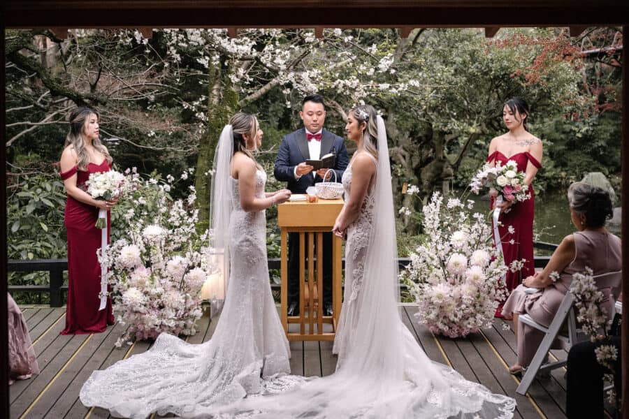 Two brides stand at a wooden lectern for their LGBTQ wedding in Japan, bridesmaids in burgundy beside them and real cherry blossom overhead.