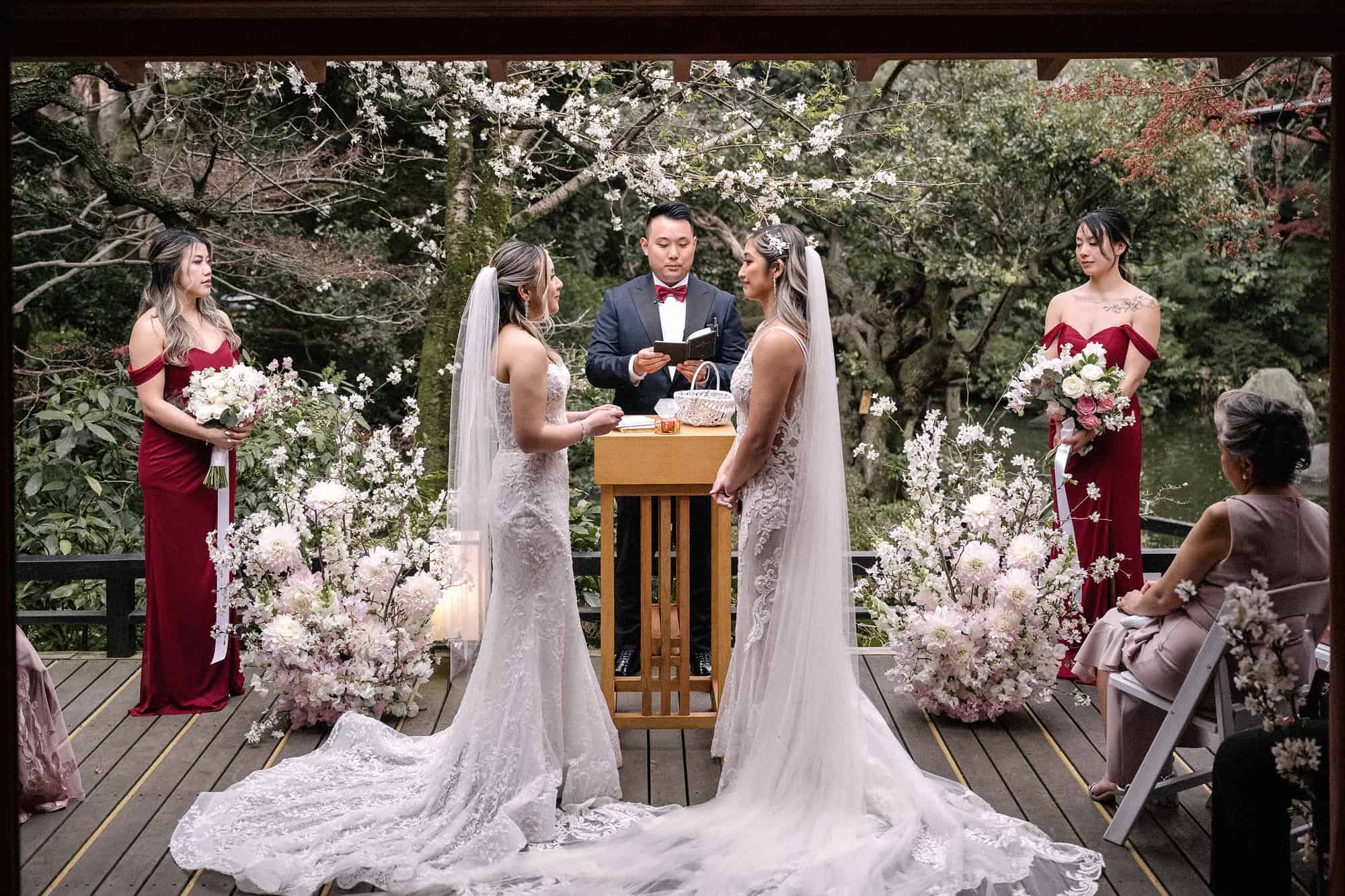 Two brides stand at a wooden lectern for their LGBTQ wedding in Japan, bridesmaids in burgundy beside them and real cherry blossom overhead.