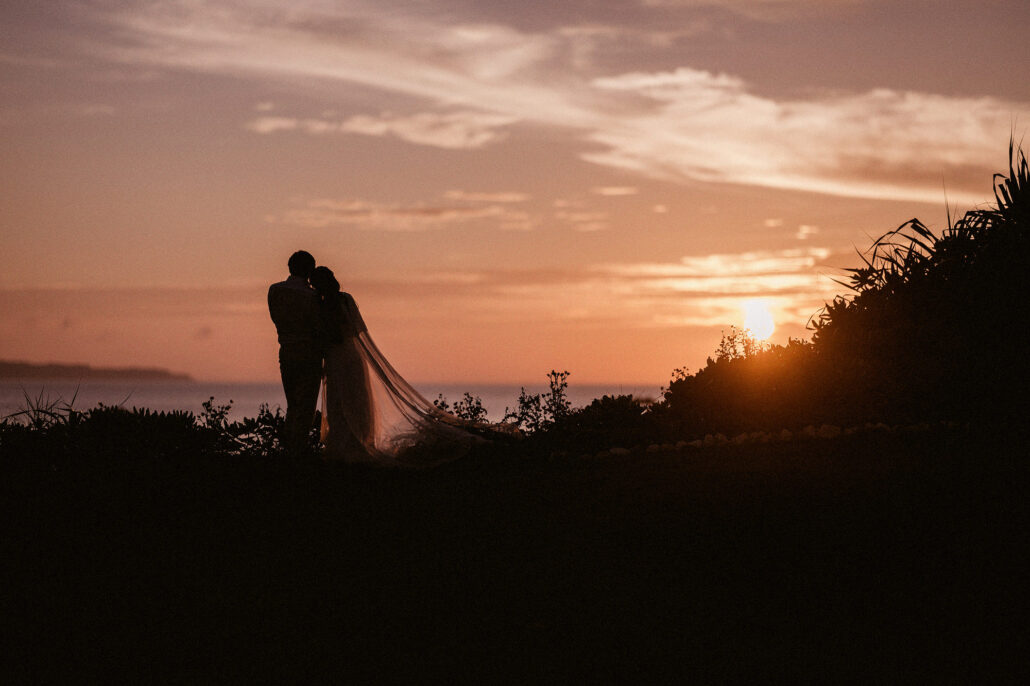 The bride and groom stand silhouetted against the sunset on a clifftop above the sea on Miyakojima.