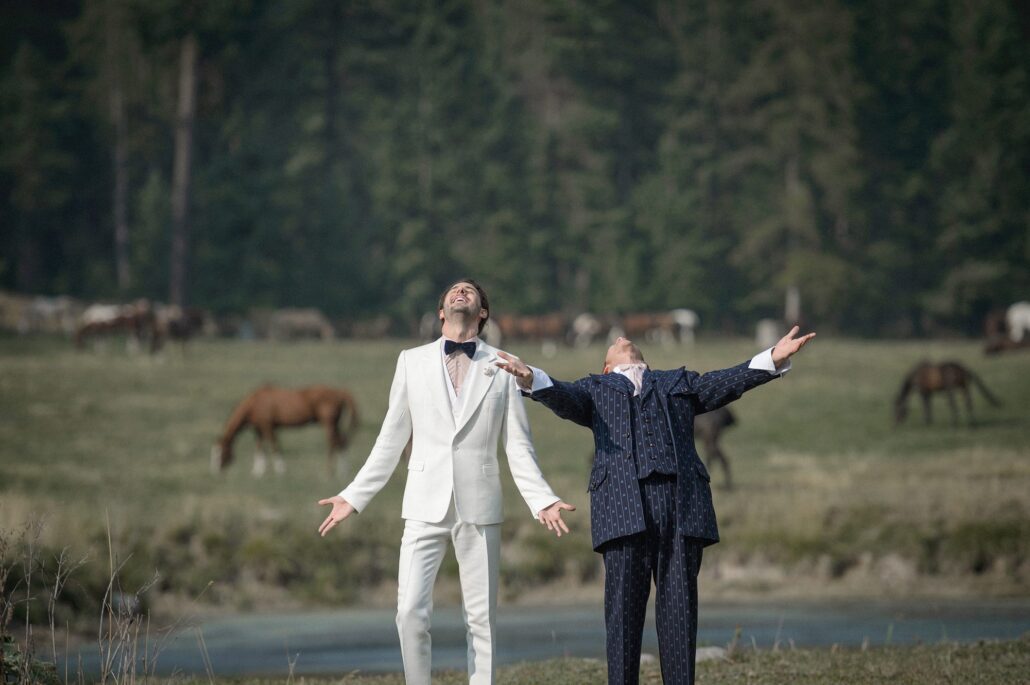 Two grooms throw their arms wide in a meadow in Montana, horses grazing behind them.