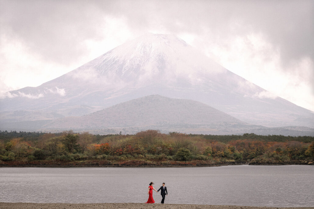 A couple stand on a lakeshore beneath Mt Fuji as cloud drifts across its slopes, the trees along the water turning for autumn.