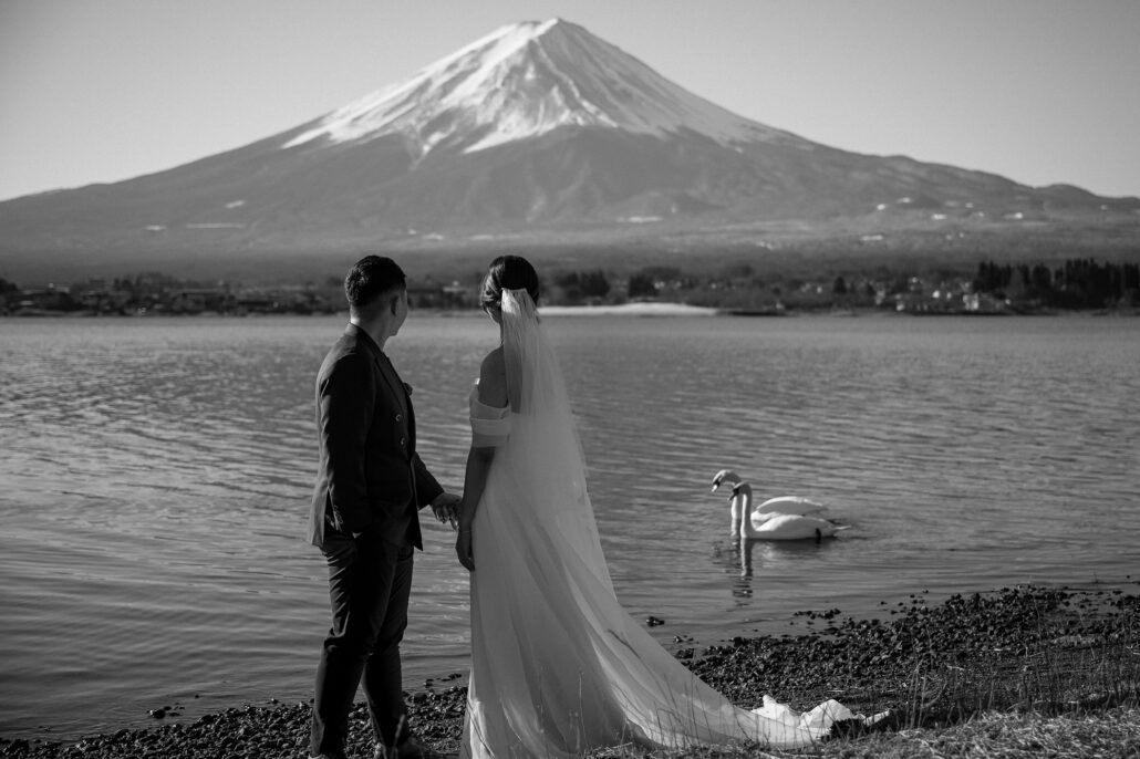 In black and white, the bride and groom stand at the edge of a lake below Mt Fuji as a swan drifts past.