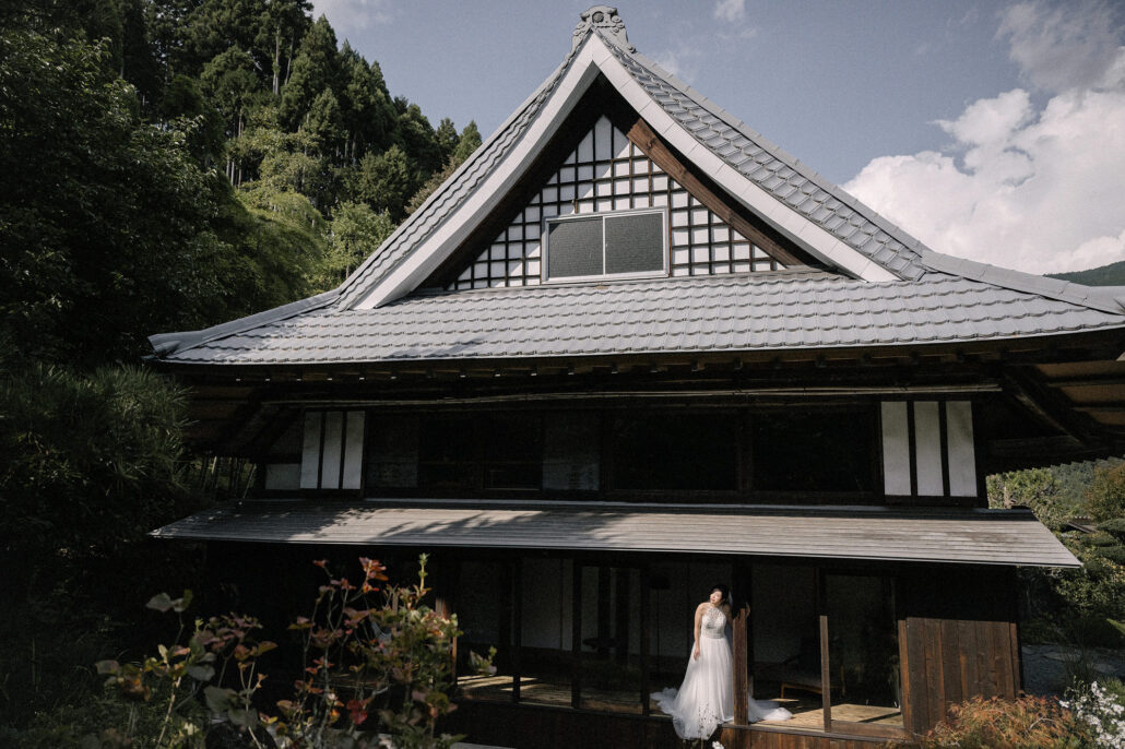 A bride stands below the steep roof of an old wooden house turned village hotel in the mountains of Japan.