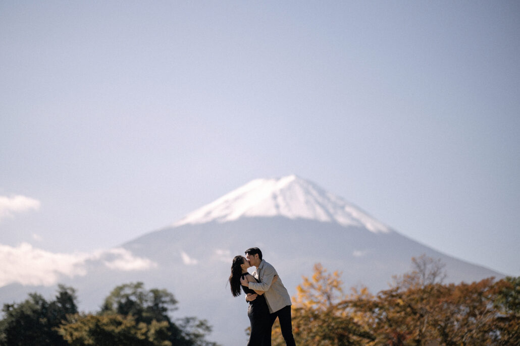 The couple kisses on the edge of an open lawn near Kawaguchiko, autumn trees turning yellow and rust below Mt Fuji.