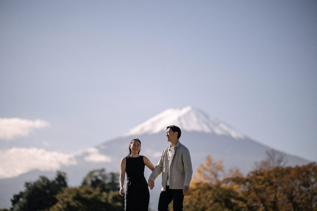 Side by side and laughing, the couple stands on an open lawn with autumn trees behind them and Mt Fuji above the clouds.