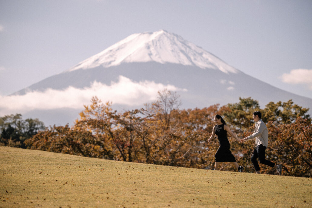 The couple runs across a golden autumn lawn near Kawaguchiko, a band of white cloud lying across the slopes of Mt Fuji.