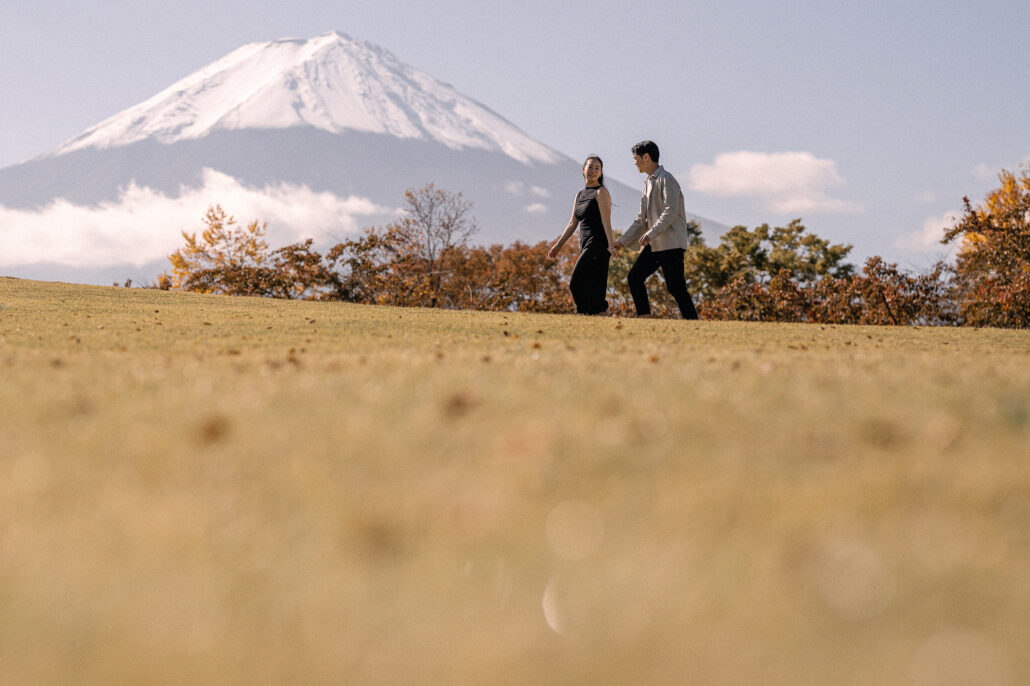 Low across the dry autumn grass, the couple walks hand in hand on the lawn as Mt Fuji and its early snow rise on the left.