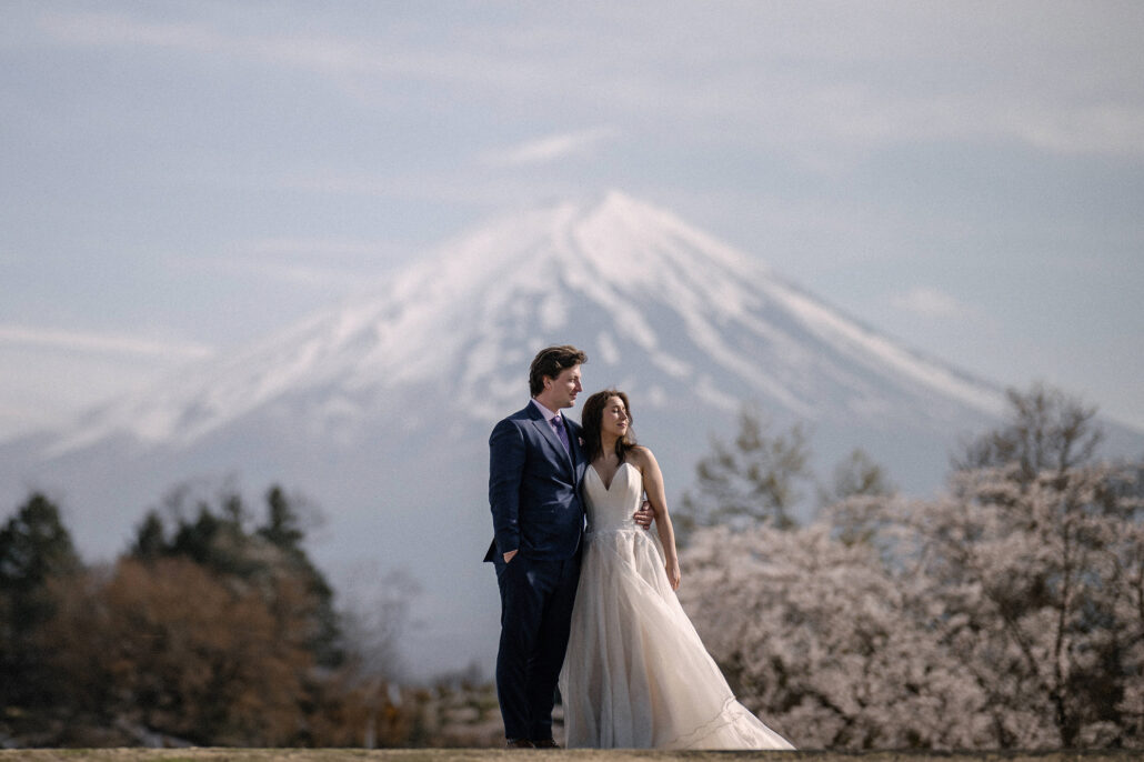 The groom and bride stand close in front of snow-capped Mt Fuji and cherry blossom trees, a Mt Fuji cherry blossom elopement portrait.