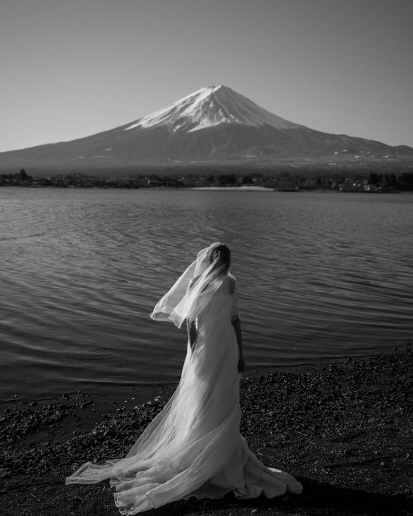 In black and white, the bride's veil lifts in the wind on a stony lakeshore, Mt Fuji white across the water.