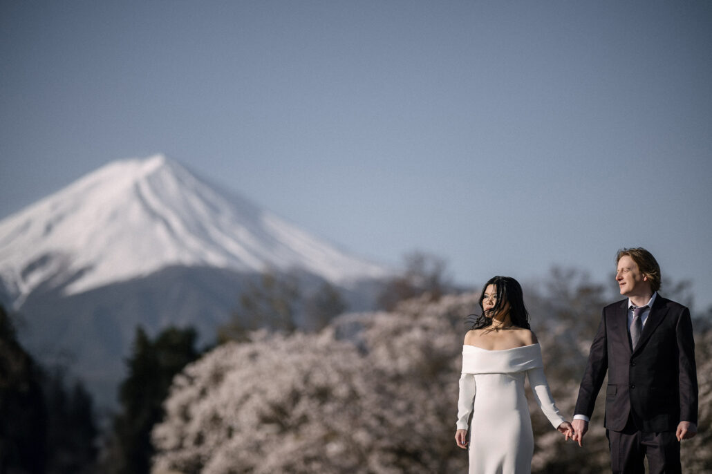 The couple hold hands in front of cherry trees in bloom, Mt Fuji rising behind them in the morning light.