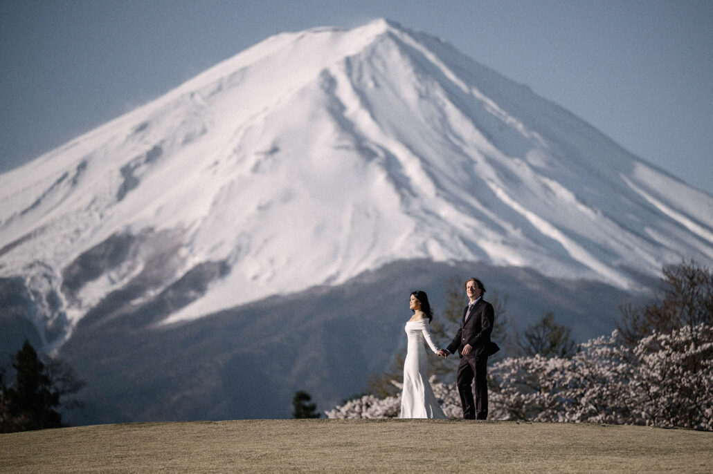 The bride and groom stand hand in hand on a lawn beneath an enormous snow-covered Mt Fuji, blossom at its foot.